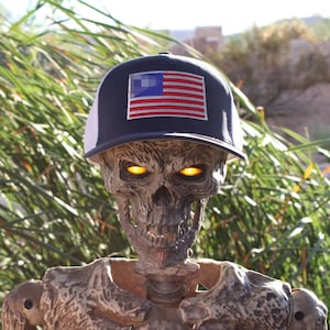 May include: A skeleton figure with glowing orange eyes wearing a navy blue and white trucker hat featuring a rectangular patch of the United States flag on the front panel. The skull has a textured, weathered appearance with deep-set eye sockets.