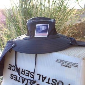 Può includere: Un cappello da sole grigio scuro con tesa larga, protezione per il collo e sottogola. La parte anteriore presenta una toppa quadrata con un design grafico blu e bianco. Il cappello poggia su una scatola bianca con la scritta United States Postal Service.