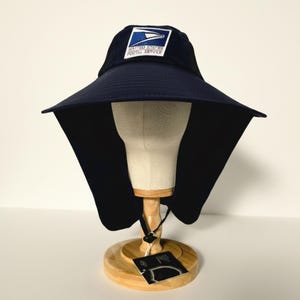 May include: A dark navy blue sun hat with a wide brim and a neck flap. The front features an embroidered patch with the United States Postal Service logo, including the eagle graphic and the text United States Postal Service in blue and red.