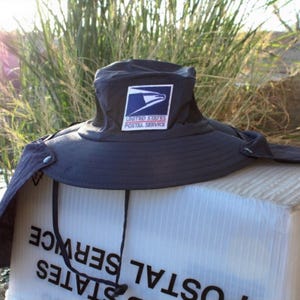 Può includere: Un cappello da sole grigio scuro a tesa larga con protezione per il collo, caratterizzato da una toppa con il logo dello United States Postal Service sul davanti. Il cappello presenta inserti laterali in rete nera e un sottogola nero sottile.