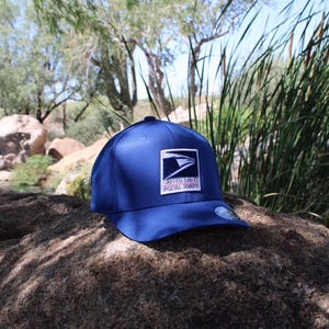 Peut inclure: Une casquette de baseball bleu roi avec un écusson rectangulaire sur le panneau avant. L'écusson affiche le logo de l'aigle du United States Postal Service en bleu foncé et blanc avec le texte UNITED STATES POSTAL SERVICE en petites lettres noires.