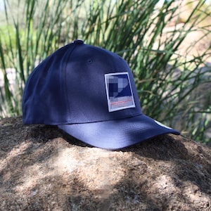 May include: A navy blue baseball cap featuring a rectangular patch on the front panel. The patch displays a dark blue square with a white graphic element and a thin red line above a white horizontal bar, set against a solid navy blue fabric.