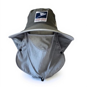 May include: A dark grey bucket hat featuring a United States Postal Service logo patch on the front. The hat includes a wide brim and a detachable light grey face and neck flap with a mesh panel, secured by black buttons and an adjustable cord.