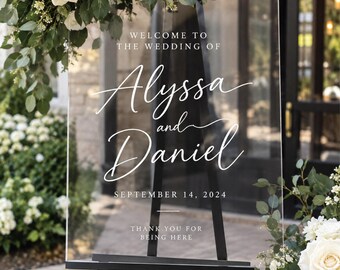 May include: A clear acrylic wedding sign featuring white printed text that reads: WELCOME TO THE WEDDING OF Alyssa and Daniel SEPTEMBER 14, 2024 THANK YOU FOR BEING HERE. The rectangular sign is displayed on a black metal easel stand.
