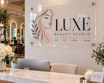 May include: A rectangular clear acrylic wall sign featuring the black text LUXE BEAUTY STUDIO, a line art illustration of a person's profile with a soft pink accent, and the words HAIR, SKIN, LASHES, BROWS, mounted with four silver standoffs.
