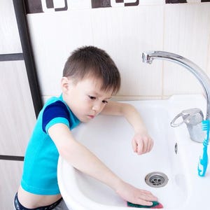 Peut inclure: Un jeune enfant en chemise bleue et manches bleu foncé nettoie un évier blanc avec une éponge verte et rouge. Un robinet chromé et une brosse à dents bleue sont également visibles.