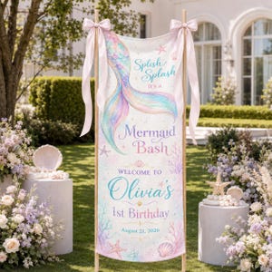 May include: A vertical banner featuring a pastel mermaid tail illustration, starfish, and seashells. The text reads: Splish Splash It's a Mermaid Bash, WELCOME TO Olivia's 1st Birthday, August 21, 2026. The banner is suspended from a wooden frame with pink ribbons.