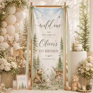 May include: A vertical fabric banner on a wooden stand featuring a forest theme with a bear, deer, and rabbit. The text reads: wild one WELCOME TO Oliver's 1ST BIRTHDAY SEPTEMBER 12, 2026. The banner is decorated with sage green ribbons at the top corners.