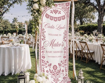 May include: A tall, vertical white fabric banner with dusty rose floral and decorative patterns. The text reads: Cielito Lindo, WELCOME TO Mateo's BABY SHOWER, NOVEMBER 14, 2026. The banner is suspended from a wooden frame and adorned with white flowers.