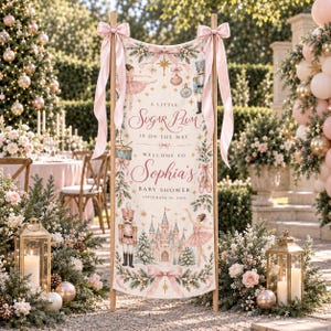 May include: A vertical banner featuring a nutcracker, ballerina, castle, and holiday ornaments with the text: A LITTLE SUGAR PLUM IS ON THE WAY WELCOME TO Sophia's BABY SHOWER SEPTEMBER 30, 2024. The banner is suspended from a wooden frame with pink ribbons.