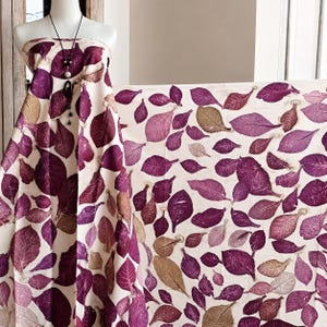 Può includere: Un capo senza spalline drappeggiato su un manichino, caratterizzato da un tessuto color crema stampato con un motivo ripetitivo di foglie viola e marrone chiaro. Una lunga collana con cordino nero, perline scure e un ciondolo centrale pende sul tessuto.