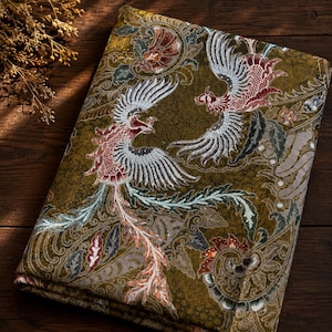 Könnte beinhalten: Ein gefaltetes Stück Stoff mit einem komplizierten Batik-Muster in Olivgrün-, Braun- und Cremetönen. Das Design zeigt zwei markante phönixartige Vögel mit detaillierten Flügeln, umgeben von wirbelnden Blumen- und Blattmotiven.
