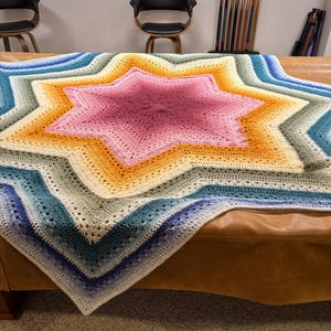 May include: A large, star-shaped crochet blanket featuring a vibrant gradient color pattern. The center begins with a deep pink, transitioning outward through shades of orange, yellow, cream, sage green, teal, and finally a dark navy blue border.