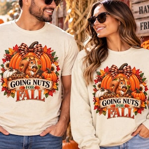 May include: A cream-coloured t-shirt and a matching sweatshirt, both featuring a graphic of a squirrel, pumpkins, sunflowers, and autumn leaves. The design includes a plaid bow and the text "GOING NUTS FOR FALL" in a decorative font.