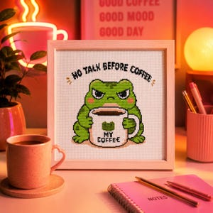 May include: A square framed cross-stitch embroidery featuring a green frog with a grumpy expression holding a white mug. The text above the frog reads NO TALK BEFORE COFFEE and the mug features a small frog icon with the text MY COFFEE.
