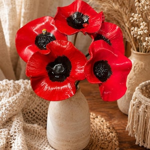 Könnte beinhalten: Ein Strauß aus fünf leuchtend roten Mohnblumen mit dunklen schwarzen Zentren, arrangiert in einer hellbeigen Keramikvase. Eine Blume ist mit einer kleinen gelb-schwarzen Bienenfigur verziert, eine andere mit einem kleinen Marienkäfer.