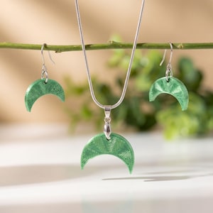 Puede incluir: Un conjunto de joyería que consta de un collar de cadena de serpiente plateada con un colgante de media luna y dos pendientes a juego. Cada pieza en forma de media luna presenta una superficie verde claro texturizada con finas líneas grabadas.