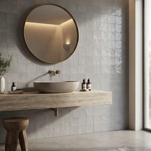 May include: A bathroom vanity area featuring a round mirror with a thin frame, a beige oval vessel sink, and a wall-mounted faucet. The sink sits on a thick, textured stone slab countertop. A small wooden stool is positioned to the left.