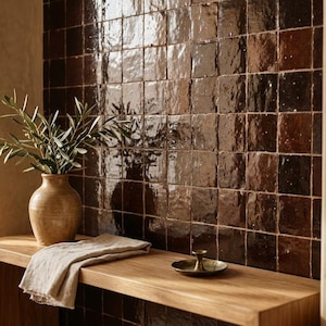 Könnte beinhalten: Eine Wand mit glänzenden, dunkelbraunen quadratischen Fliesen mit unebener, reflektierender Oberfläche. Eine hellbraune Keramikvase mit grünen Olivenzweigen steht neben einem gefalteten beigen Leinentuch und einer kleinen Messingschale.