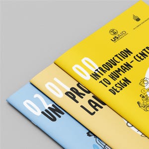 May include: Three colorful booklets with the titles "Introduction to Human-Centered Design" and "Un Pro Lan" are stacked diagonally. The top booklet is yellow, the middle is beige, and the bottom is light blue. The booklets have white text and illustrations.