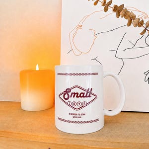 Puede incluir: Taza de cerámica blanca con el texto "Small Joys" en un diseño de rombo burdeos. La taza también presenta el texto "A Reason to Stay Since 2026". Una vela naranja encendida y un dibujo lineal están en el fondo.