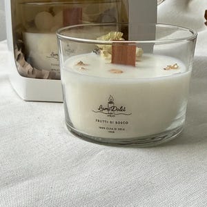 Op de afbeelding: Een glazen kaars met een houten lont en witte was, versierd met gedroogde bloemen en goudvlokken. De kaars zit in een transparante glazen houder, met de tekst "Lumi Dolci ATELIER FRUTTI DI BOSCO 100% CERA DI SOIA 140GR".
