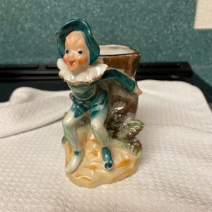 Könnte beinhalten: Vintage Keramikfigur eines lächelnden Elfen in teal- und weißer Kleidung. Der Elf lehnt an einem braunen Baumstumpf mit einer kleinen Öffnung. Der Sockel ist hellbeige mit grünen und braunen Akzenten.