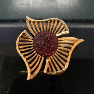 May include: A gold-toned metal flower brooch with a central cluster of red rhinestones. The brooch features three stylized petals and radiating lines, creating a floral design. The brooch is set against a dark background.