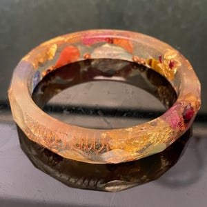 Könnte beinhalten: Ein klarer Harz-Armreif mit eingebettetem Blattgold und bunten getrockneten Blumen. Das Armband hat ein facettiertes Design und einen Durchmesser von etwa 6,35 cm.