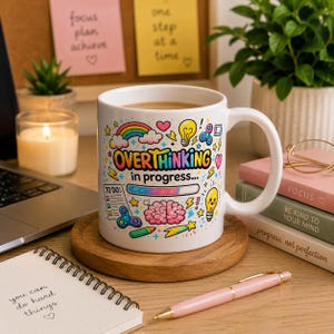 Può includere: Una tazza in ceramica bianca con un design grafico colorato che riporta il testo "OVERTHINKING in progress..." sopra una barra di caricamento. L'illustrazione include un cervello rosa, una lampadina, un arcobaleno e una lista di cose da fare.