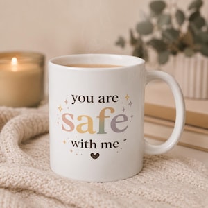 Può includere: Tazza in ceramica bianca con la scritta "you are safe with me" in colori pastello e un simbolo a forma di cuore. La tazza ha un manico curvo ed è riempita con una bevanda calda. La tazza è appoggiata su una coperta di maglia color crema.