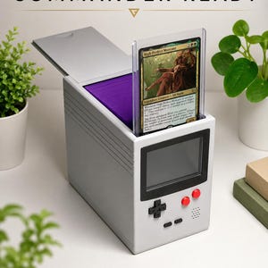 May include: A gray card storage box with a retro video game console design, holding a Magic: The Gathering card. The box has a flip-top lid, purple interior, and red buttons. Text on the box reads "COMMANDER READY" and "DISPLAY YOUR COMMANDER • STORE 100+ DOUBLE-SLEEVED CARDS."