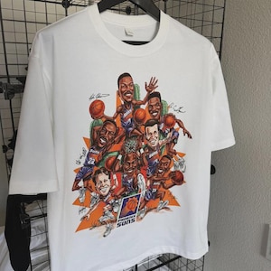 May include: White t-shirt featuring a cartoon illustration of Phoenix Suns basketball players. The graphic includes the team logo and player signatures. The shirt is hanging on a black hanger against a wire rack.