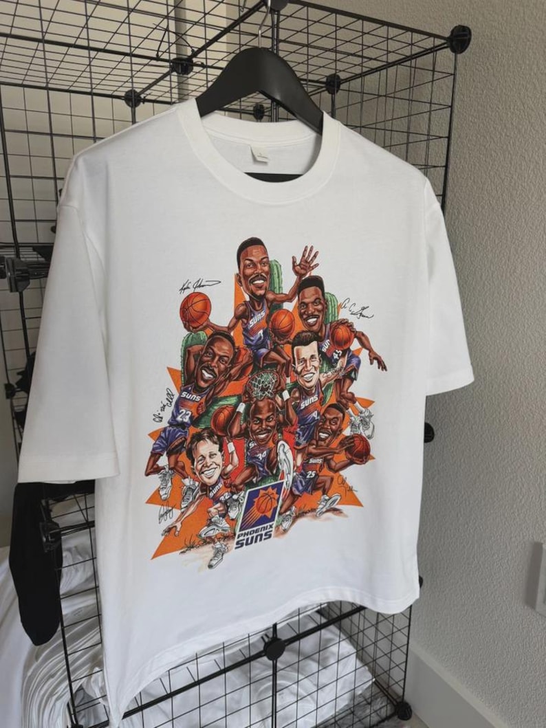 May include: White t-shirt featuring a cartoon illustration of Phoenix Suns basketball players. The graphic includes the team logo and player signatures. The shirt is hanging on a black hanger against a wire rack.