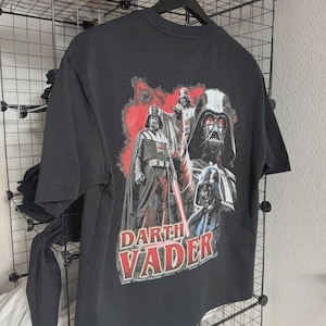 May include: Black t-shirt featuring a graphic of Darth Vader from Star Wars. The design includes multiple images of Darth Vader with a red and white color scheme. The text "Darth Vader" is printed in red and white.