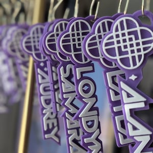 May include: A row of purple and white custom keychains hanging from a gold metal rack. Each keychain features a clover-shaped icon at the top and a name printed in white capital letters, including the names Audrey, Londyn, Samara, and Alana.