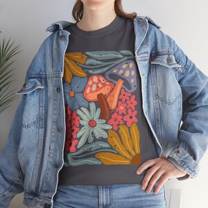 Könnte beinhalten: Dunkelgraues T-Shirt mit einem farbenfrohen Blumen- und Pilzdesign. Die Grafik zeigt orange, rote, blaue und gelbe Blumen sowie orange und lila Pilze. Das Shirt wird mit einer Jeansjacke getragen.