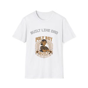 Peut inclure: Un t-shirt blanc à manches courtes avec une impression graphique sur la poitrine. Le texte indique BUILT LIKE DAD en haut et POLY BOY BUILT DIFFERENT en bas. L'illustration centrale représente une personne aux bras croisés dans un cadre.