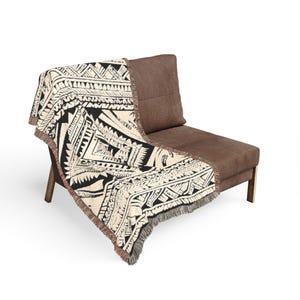 Peut inclure: Un plaid tissé présentant un motif géométrique contrasté noir et crème avec des motifs complexes d'inspiration tribale. Le textile rectangulaire possède une bordure à franges sur le bas et est drapé sur une chaise rembourrée marron.