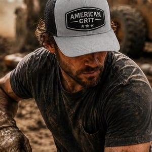 May include: Gray and black trucker hat with the text "AMERICAN GRIT" in a black and white patch. The hat has a curved brim and a mesh back. The person in the image is wearing a dark gray t-shirt.