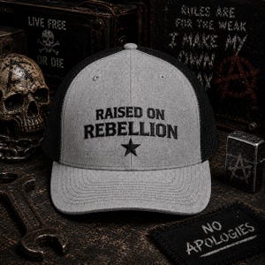 May include: A gray trucker hat with a black mesh back. The front of the hat features the embroidered text "RAISED ON REBELLION" above a black star. Additional text and imagery are visible in the background.