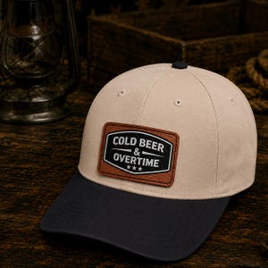 Op de afbeelding: Beige baseballpet met een zwarte klep. De pet heeft een bruin leren patch met de witte tekst "COLD BEER & OVERTIME" en drie sterren. Een casual accessoire.
