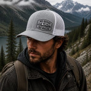 May include: Gray trucker hat with a white mesh back and a curved brim. The hat features a rectangular patch with the text "NO ROADS NO RULES" above a mountain graphic. The hat is worn outdoors with a mountain backdrop.