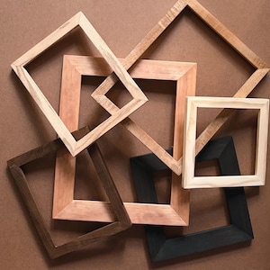 Può includere: Una collezione di cinque cornici in legno disposte in un motivo sovrapposto. Le cornici presentano varie finiture, tra cui legno naturale chiaro, marrone medio, marrone scuro e nero, ciascuna con un design a bordo semplice e piatto.