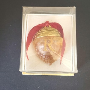 May include: An acorn-shaped ornament featuring a translucent amber-colored body with a textured surface and a gold-toned cap. The letter R is printed in gold on the front, and a dark red ribbon is tied at the top, all encased in a clear box.