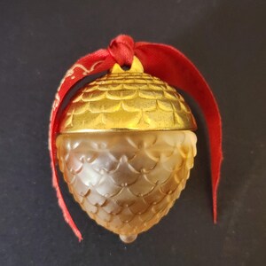 May include: An acorn-shaped ornament featuring a metallic gold cap with a textured scale pattern and a frosted amber glass base. A red satin ribbon is tied through the top loop, with the fabric ends draping down on both sides of the object.