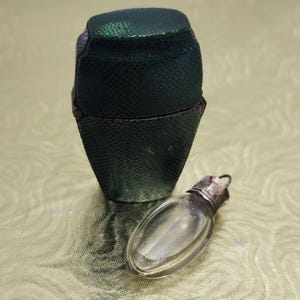 May include: A dark green, textured, hexagonal-shaped case with a hinged lid sits upright. Beside it lies a small, clear, oval-shaped glass vial featuring a metallic silver-toned cap with a small loop at the top for hanging or attachment purposes.