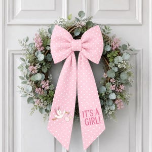 Op de afbeelding: Een decoratieve krans met groene eucalyptusbladeren en kleine roze bloemen, versierd met een grote roze strik met witte stippen. Eén uiteinde van de strik toont een ooievaar met een baby, het andere uiteinde bevat de tekst IT'S A GIRL!