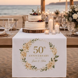 Peut inclure: Un chemin de table blanc orné d'une couronne florale dorée avec le texte 50th ANNIVERSARY EST. 1974 et les prénoms Jessie & Adam imprimés au centre, disposé sur une table en bois préparée pour une célébration en extérieur avec un gâteau.