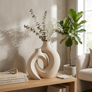 Peut inclure: Deux vases en céramique beige avec un design entrelacé unique, contenant de l'eucalyptus et des fleurs blanches. Les vases sont posés sur une table en bois clair, avec des livres et un petit bougeoir doré. Une plante en pot et un fauteuil crème sont en arrière-plan.